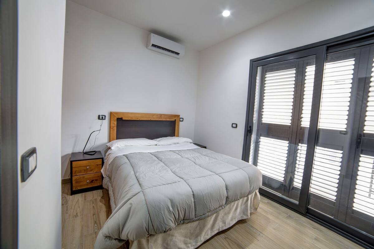 A bedroom with a bed, a nightstand, an air conditioning unit, and a sliding glass door.