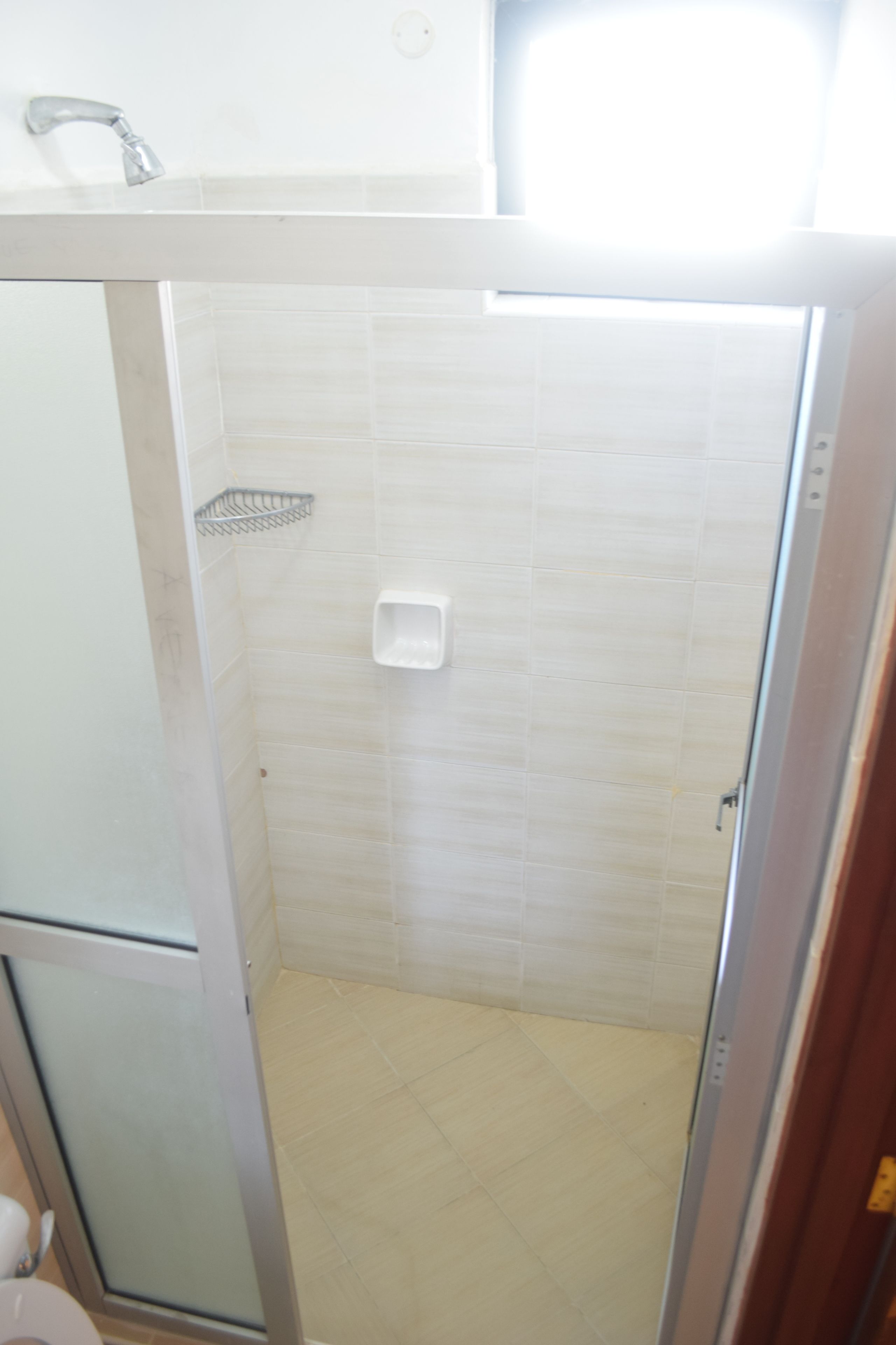 Een klein, afgesloten douchegedeelte met een witte betegelde wand, een metalen douchebak en een witte zeepdispenser.