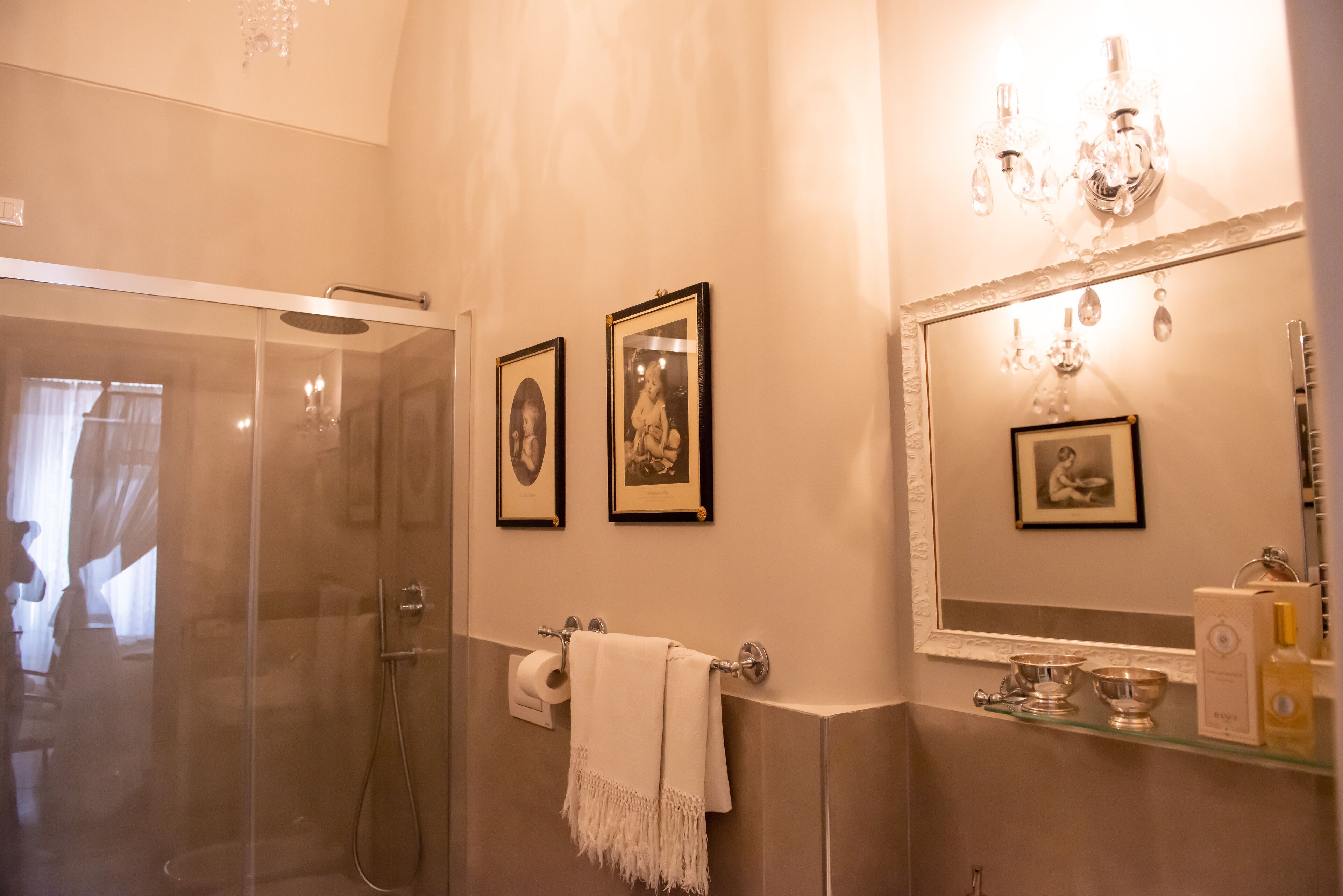 Ein Badezimmer mit Duschkabine aus Glas, einem großen Spiegel mit einer Chandelier und gerahmten Bildern an der Wand.