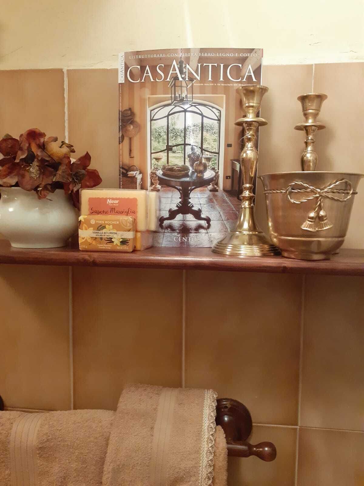 Een tijdschrift met de titel 'CASANTICA' op een plank, een vaas met gedroogde bloemen, een doos 'Near Sapore Marisciglia' vanillekoekjes, een messing kandelaar en een zilveren kom met een touw erom.