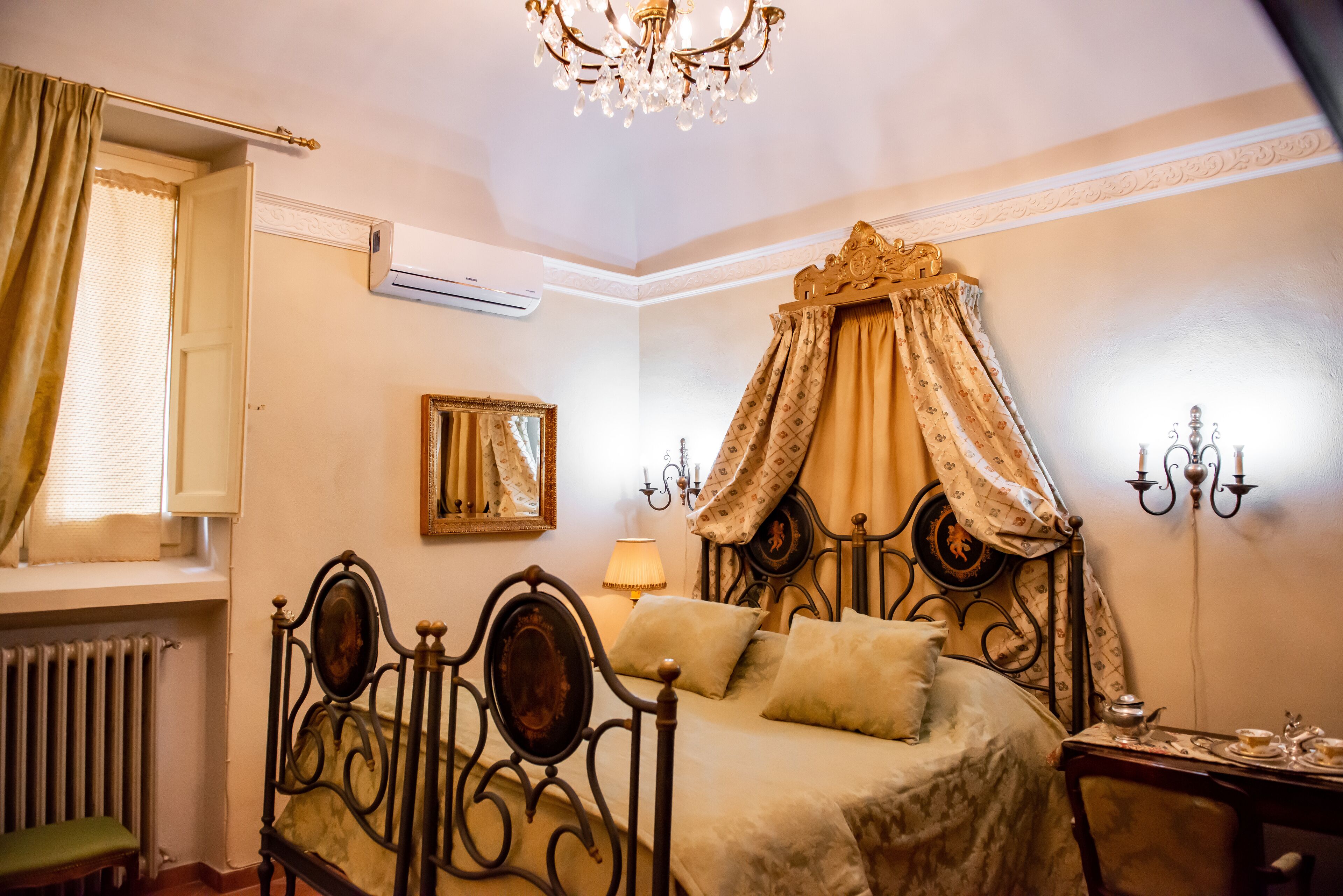 A bedroom with a four-poster bed, a chandelier, a bedside table with a lamp, and a radiator.