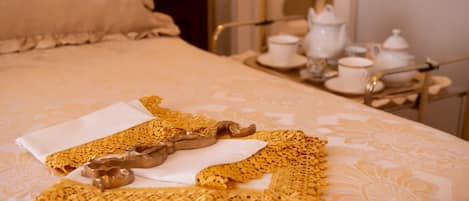 A neatly made bed with a lace bedspread, a gold decorative pillow, and a tray with tea cups and a teapot.