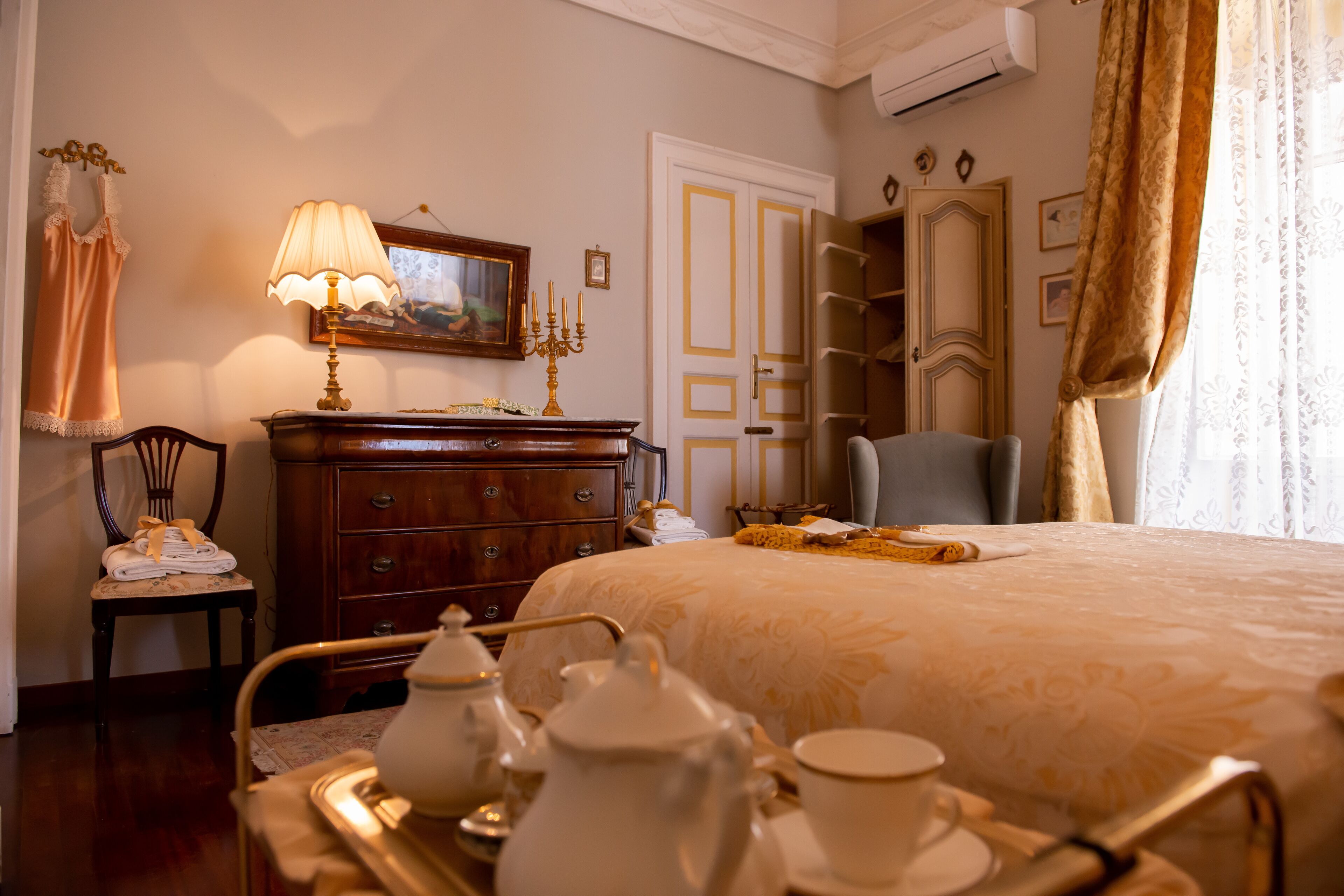 A hotel room with a bed, a wooden dresser, a chair, a table with tea set, and a painting on the wall.
