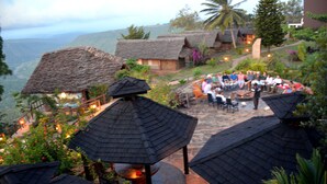 A group of people gathered around a fire pit in a tropical setting with thatched-roof huts and lush greenery.