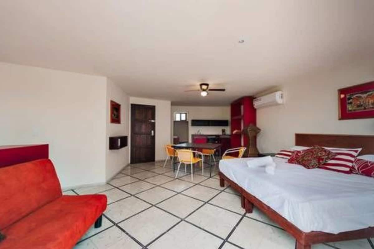 Una habitación con una cama, un sofá rojo, una mesa de comedor con sillas y una zona de cocina.