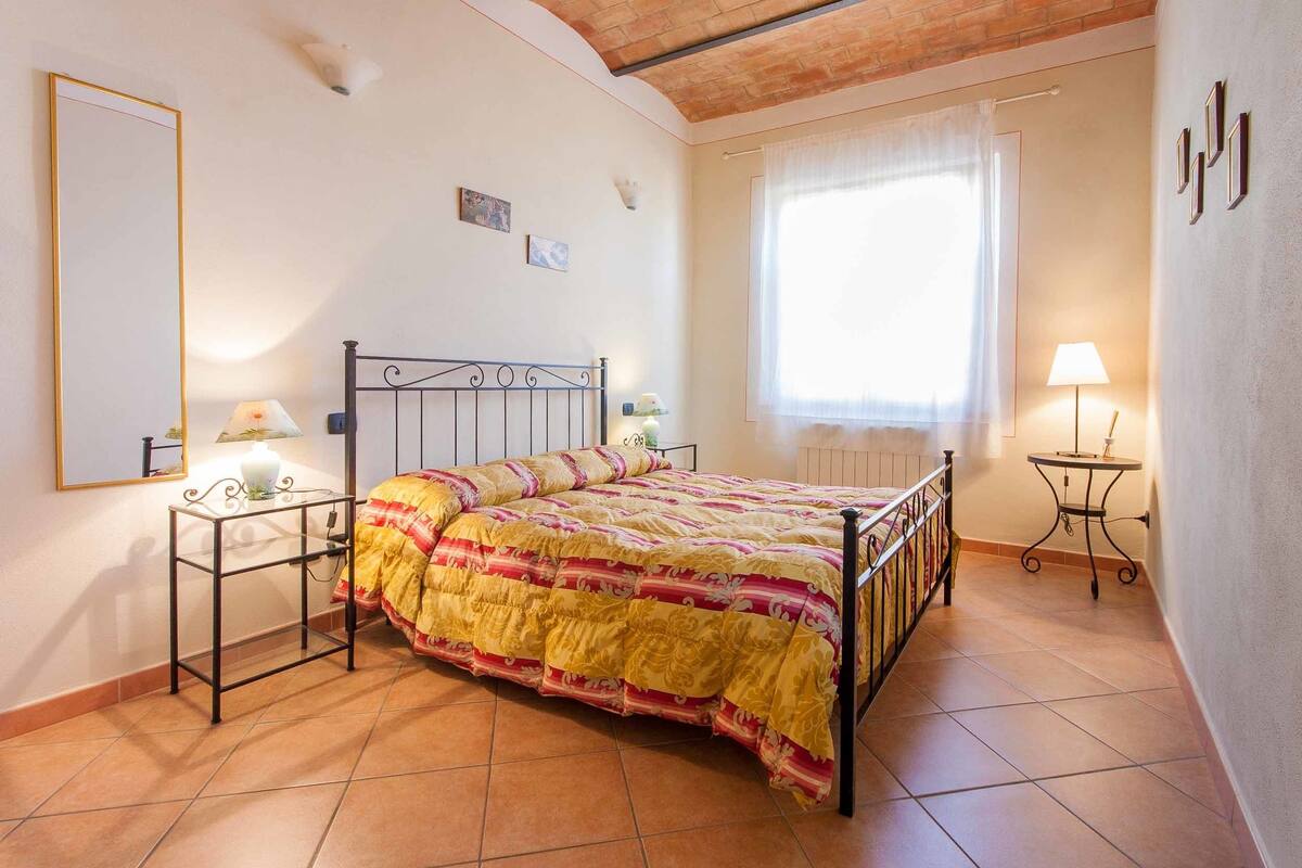 Ein Schlafzimmer mit Bett, Nachttischen, einem Fenster mit Vorhängen und einem Fliesenboden.