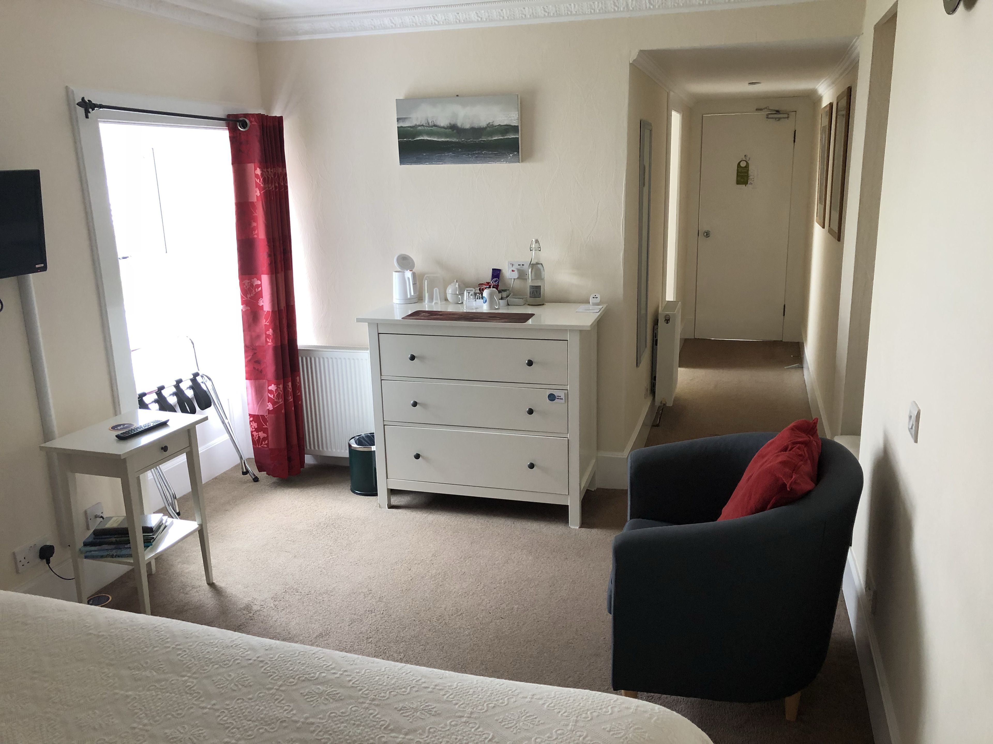 A hotel room with a bed, a desk with a computer, a chair, a dresser, and a window with red curtains.