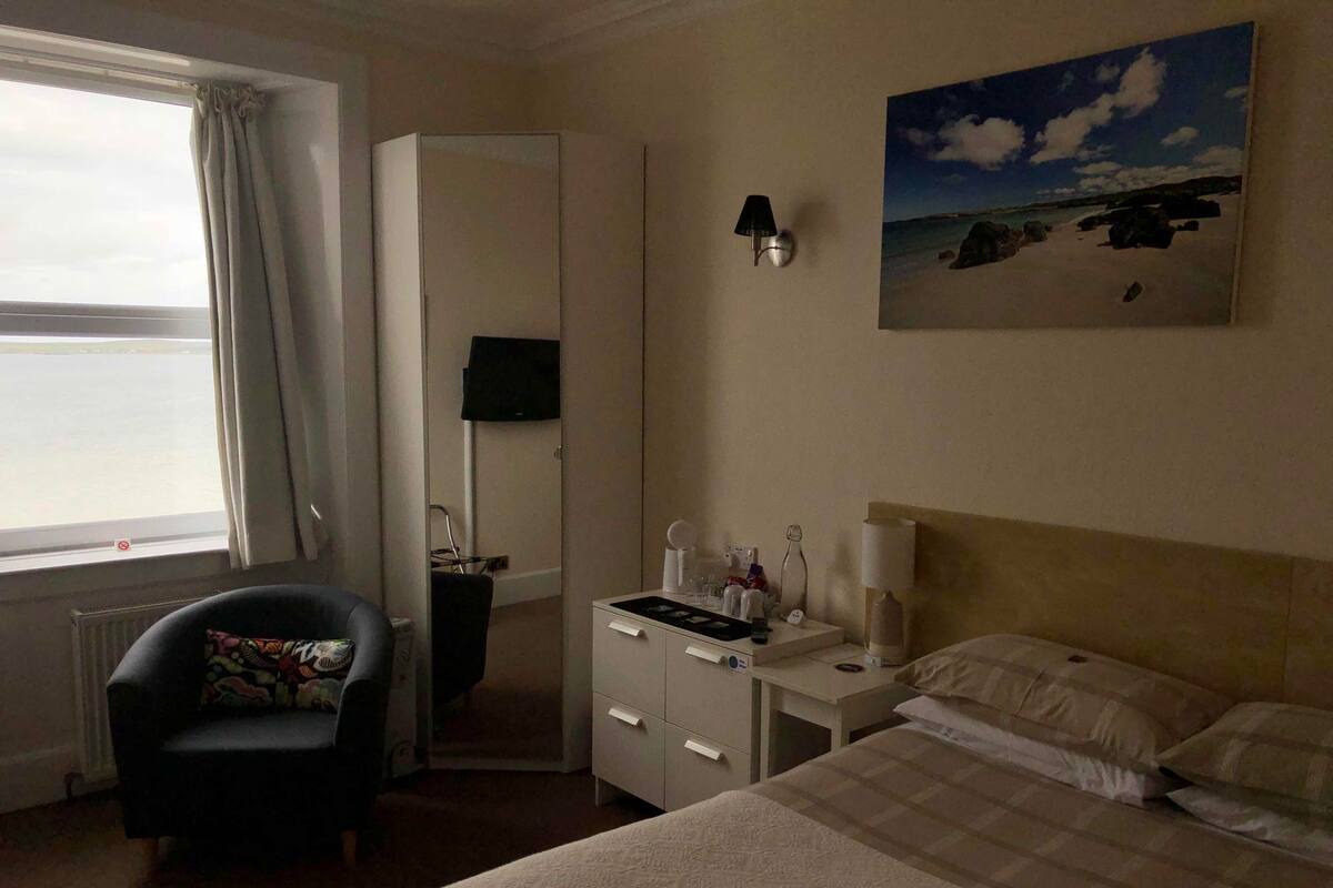 A hotel room with a bed, a chair, a desk with a lamp, a painting on the wall, and a window with curtains.