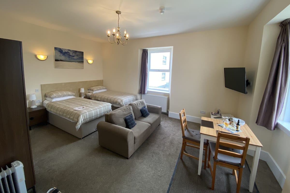 A hotel room with two beds, a sofa, a dining table with chairs, a chandelier, a painting on the wall, and a window with curtains.