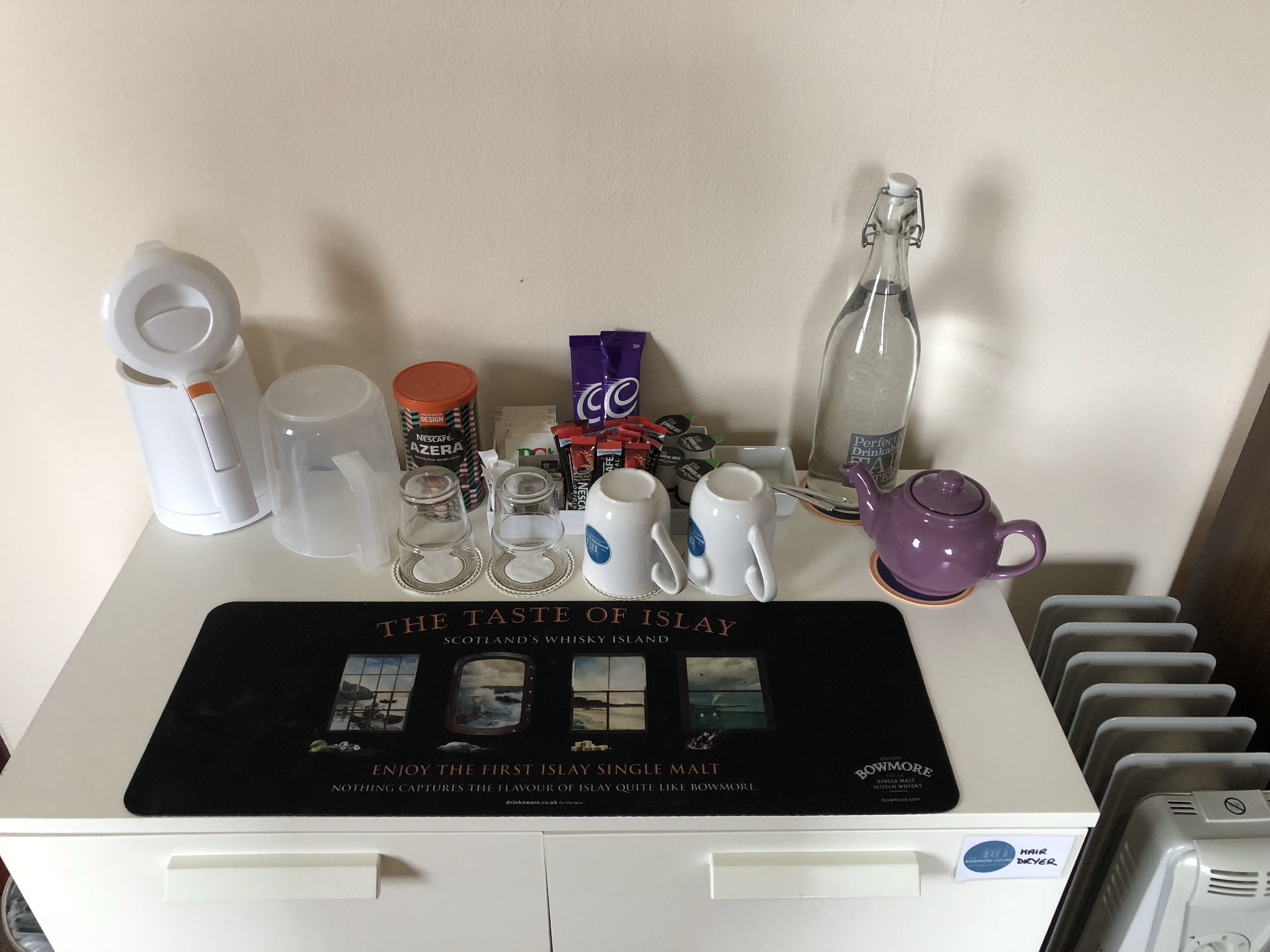A table with a whisky advertisement, various mugs, a teapot, and a hairdryer.