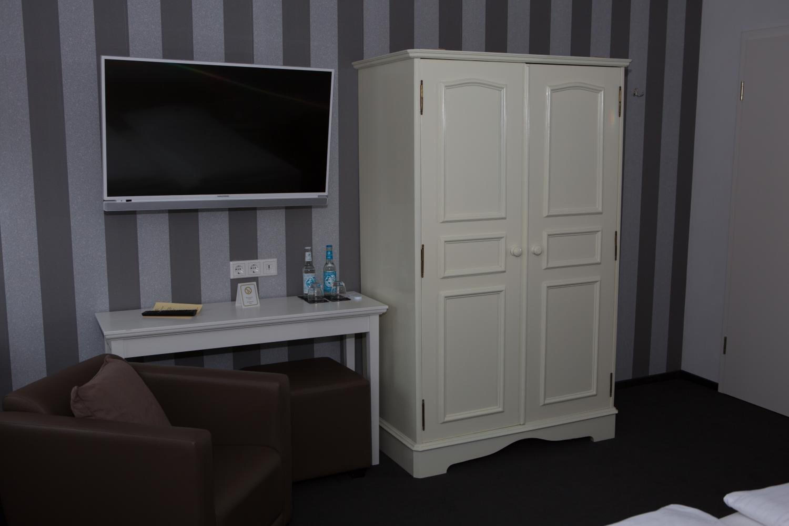 A room with a television mounted on the wall, a white cabinet, a small table with a remote control, and two brown chairs.