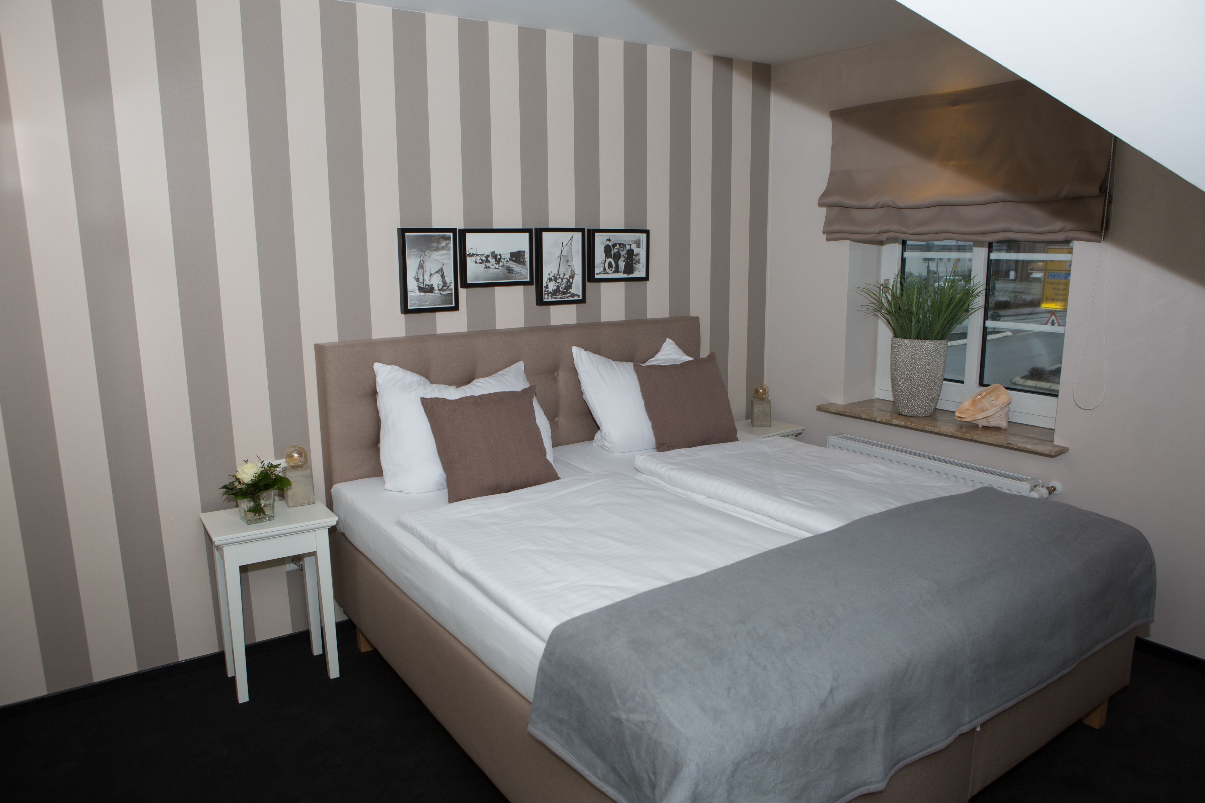 A bedroom with a large bed, striped wallpaper, a small bedside table with a vase, and a window with a plant.