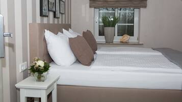 A bedroom with a bed, pillows, a bedside table with a vase of flowers, a window with a plant, and a wall with framed pictures.