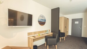 Habitación de hotel con televisión de pantalla plana, un escritorio con silla, un pequeño cubo de basura y una puerta.