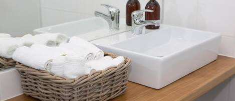 Un baño con un lavabo blanco, dos grifos y una cesta de mimbre llena de toallas blancas.