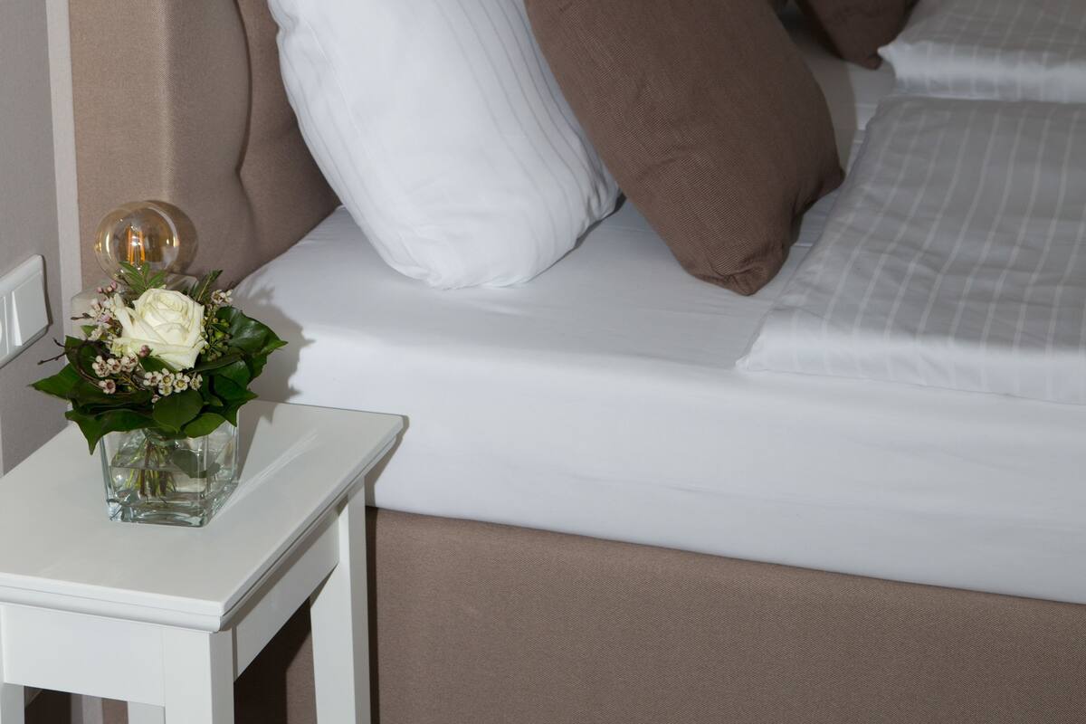 Una cama bien hecha con almohadas blancas y marrones, una mesita de noche con un florero y cuadros en la pared.