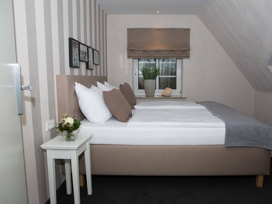 A bedroom with a bed, a bedside table with a vase of flowers, a window with a plant, and a wall with striped wallpaper.