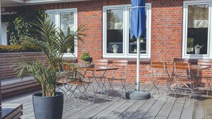 Una terraza de madera con asientos exteriores, una jardinera grande con una planta de palmera y un edificio de ladrillo al fondo.