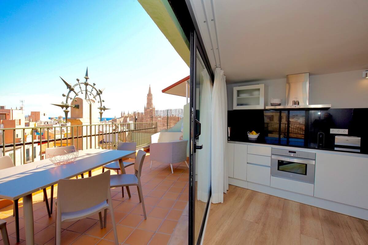 Eine moderne Wohnung mit Balkon, einer Küche mit Einbauofen und Blick auf eine Stadtlandschaft, inklusive eines Kirchturms.