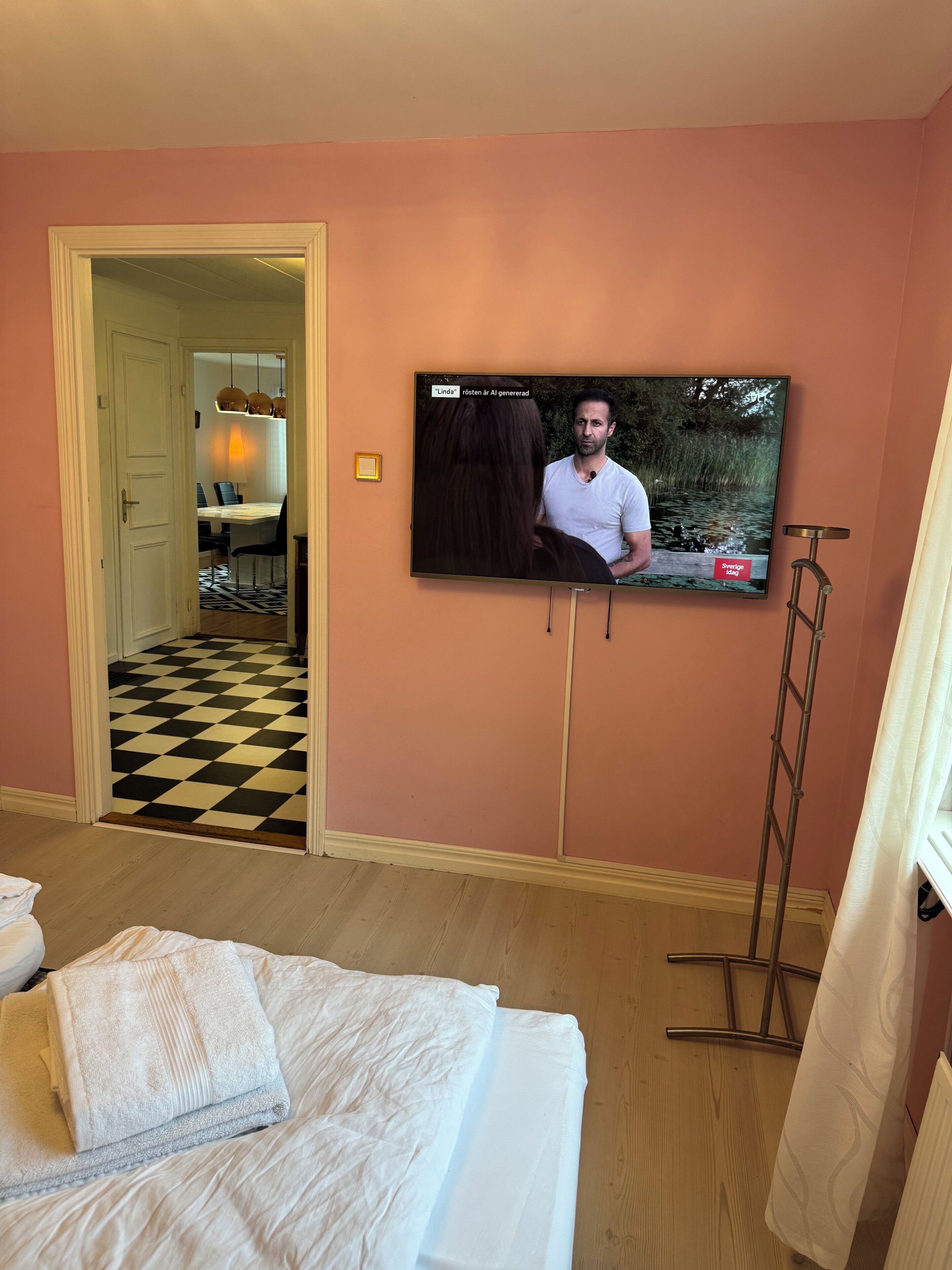 Une chambre d’hôtel avec un lit, un téléviseur fixé au mur, une porte donnant sur un coin repas et un sol à carreaux.