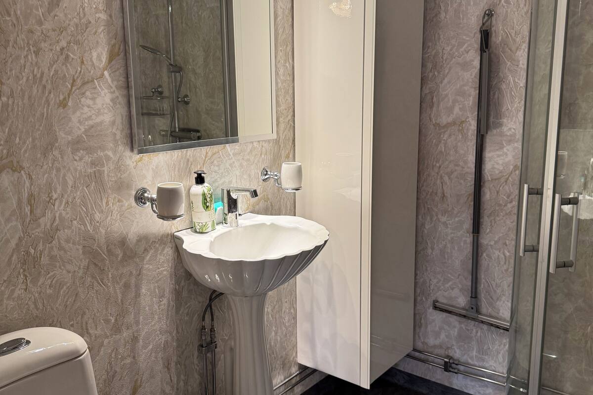 A modern bathroom with a marble wall, a pedestal sink, a toilet, a glass shower enclosure, and a vanity cabinet.