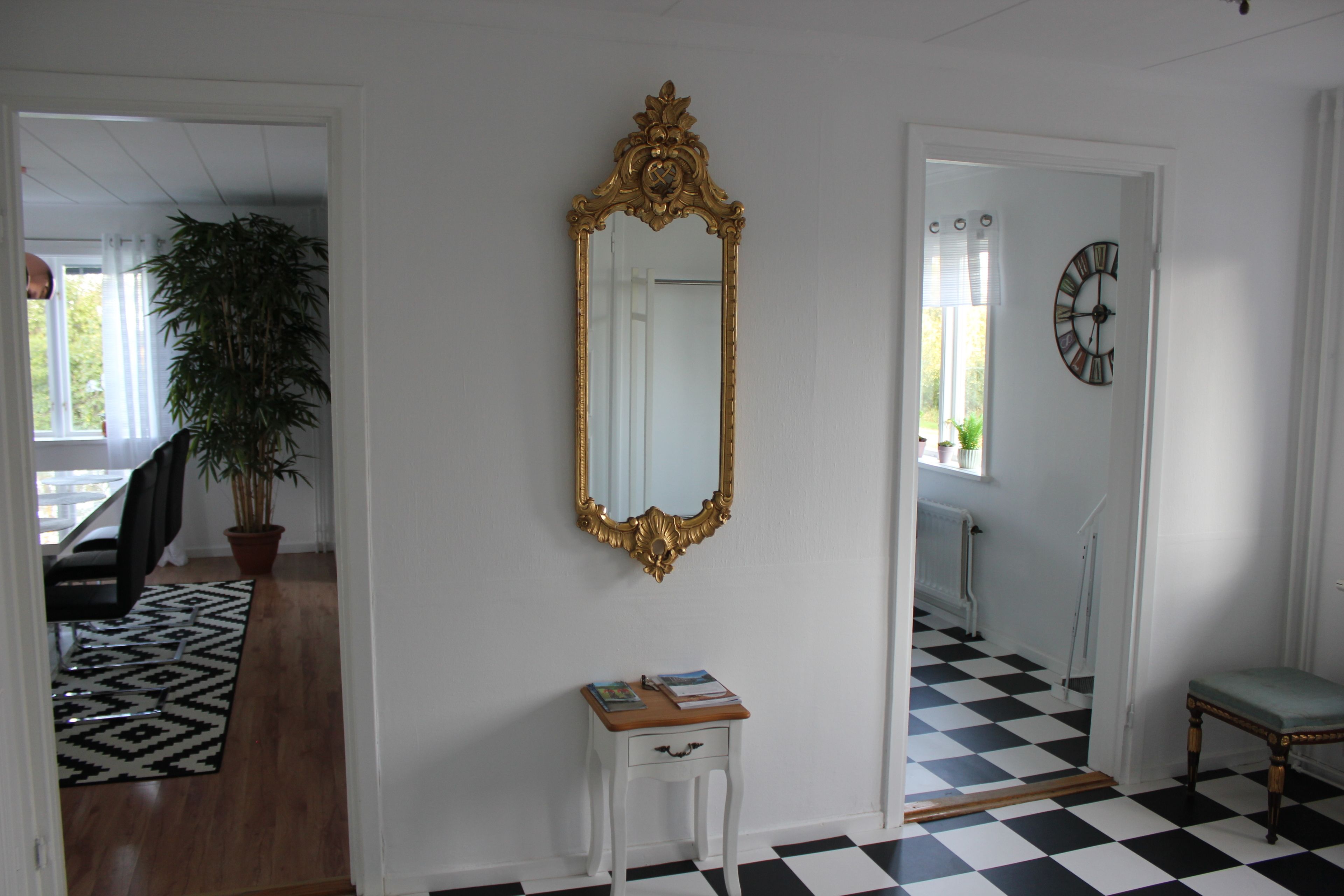 Une pièce avec un sol à carreaux, un mur blanc orné d’un miroir à cadre doré, une petite table blanche sur laquelle repose un livre, et une plante en pot.