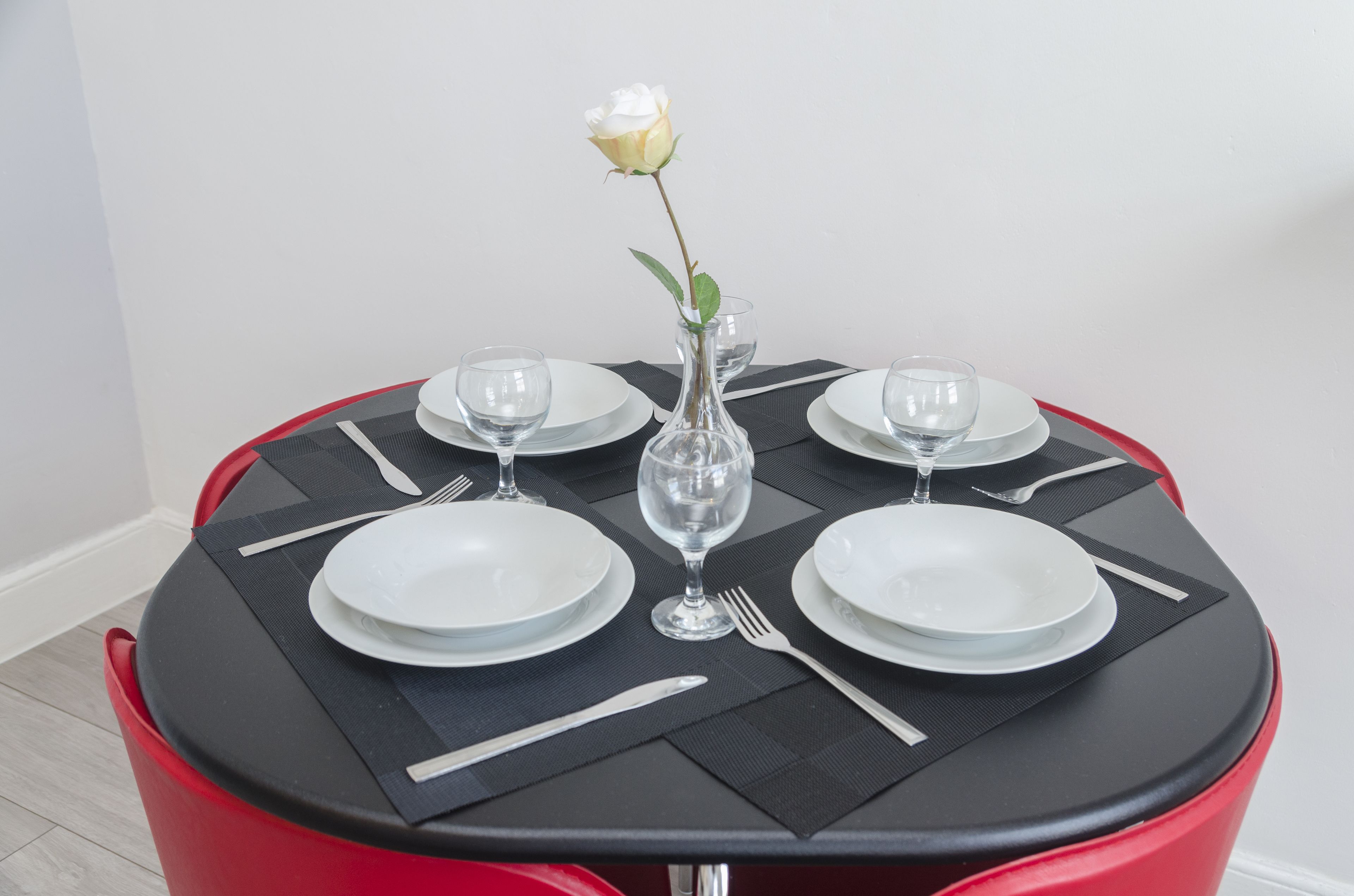 Uma mesa de jantar redonda com jogos americanos pretos, pratos brancos e copos de vidro transparente. Uma única rosa branca está colocada em um vaso transparente no centro.