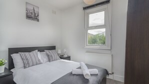 Et moderne soveværelse med en stor seng, et natbord med en lampe, et billede på væggen og et vindue med udsigt over grøn vegetation.