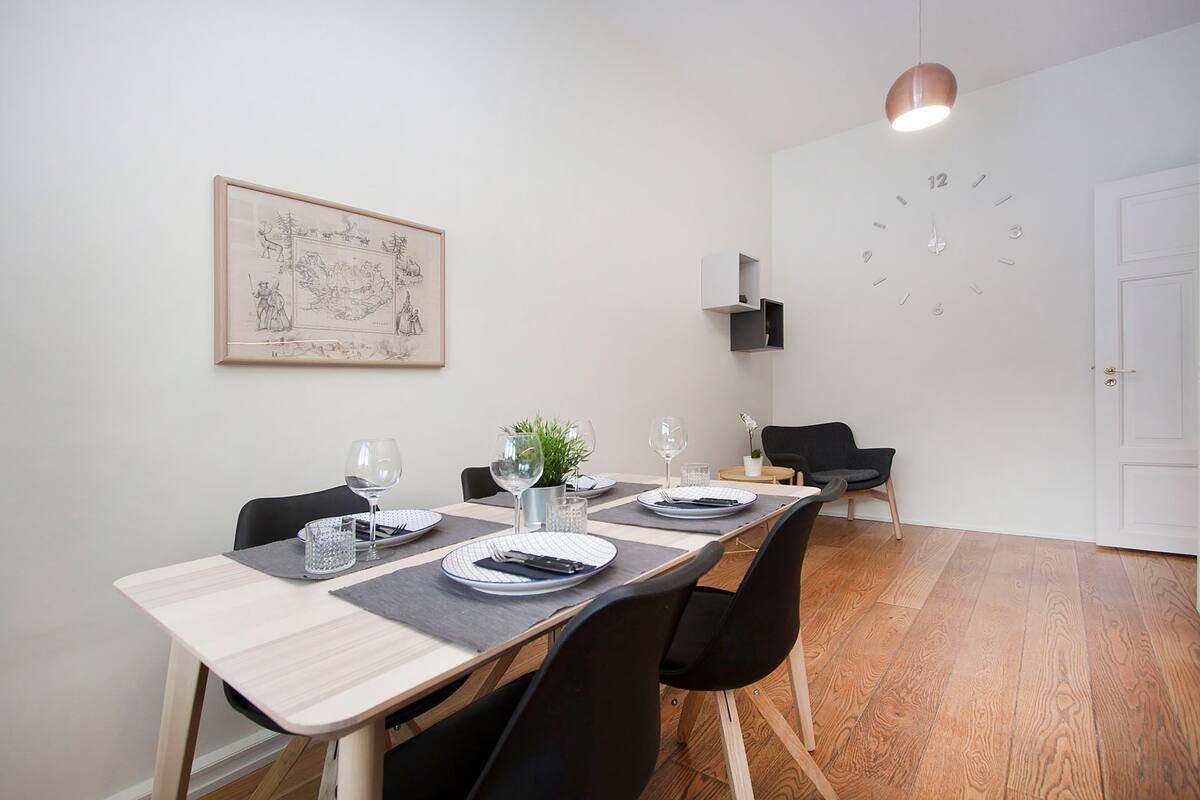 A dining area with a long table set for four, chairs, a clock, a framed artwork, and a hanging light fixture.
