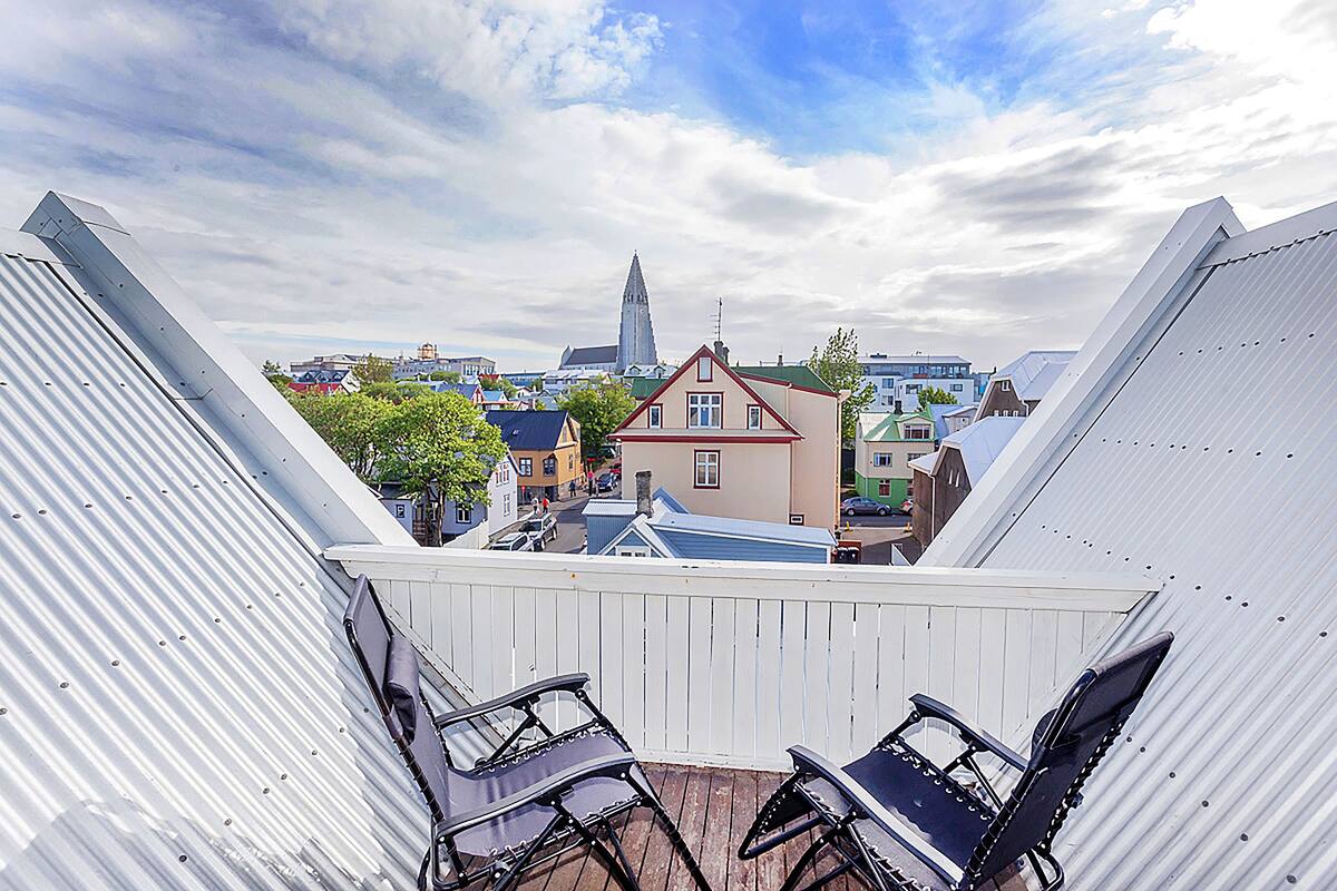 A view from a balcony with metal railings, overlooking a town with various buildings, including a church with a tall spire.