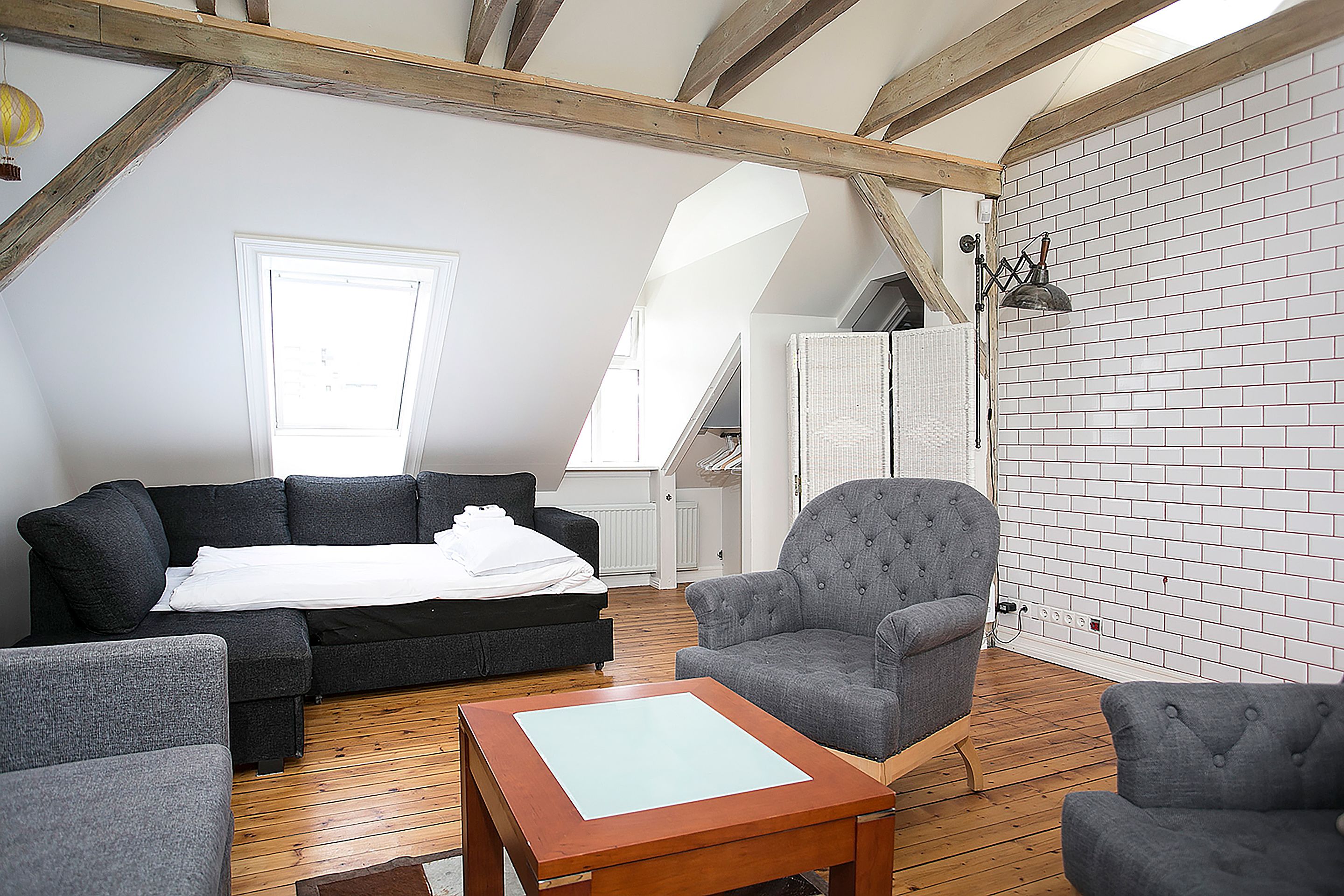 Quarto estilo loft com piso de madeira, sofá preto, poltrona cinza e uma pequena mesa de madeira.