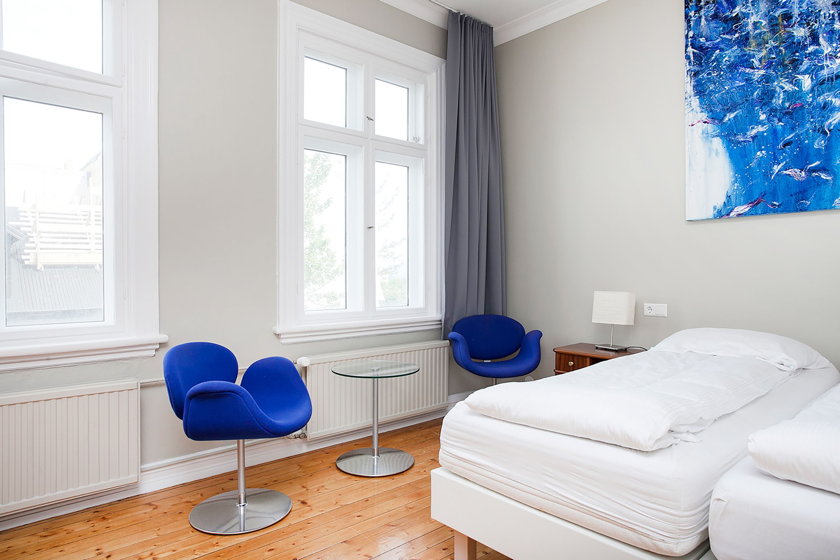 Une chambre d’hôtel avec un lit, deux chaises bleues, une petite table et un grand tableau abstrait accroché au mur.