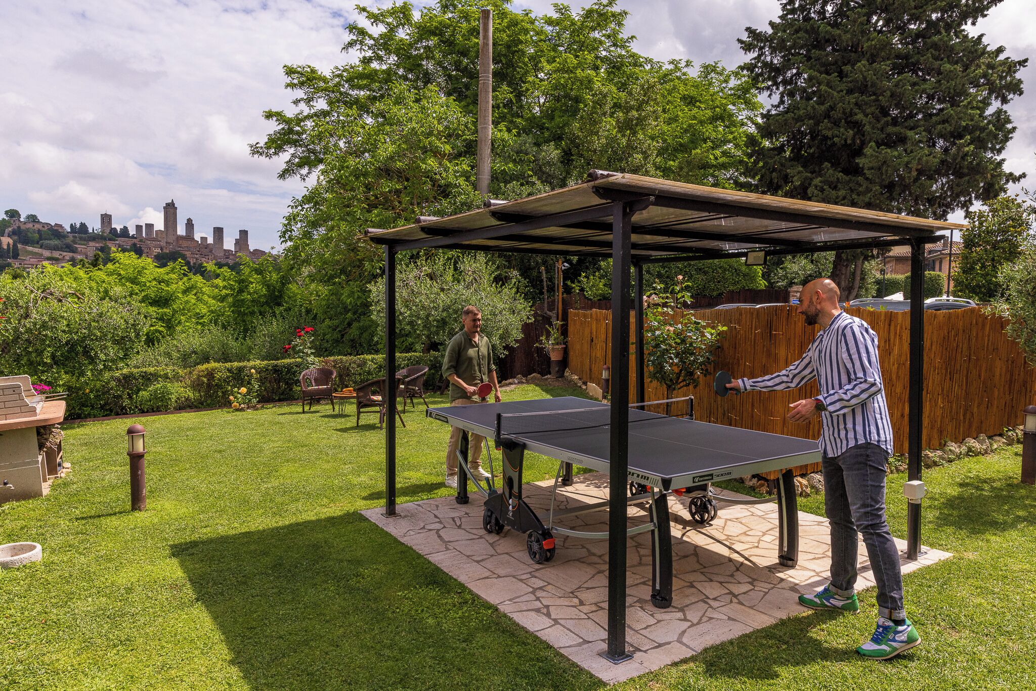 An outdoor ping pong table set up on a stone patio with a pergola overhead.
