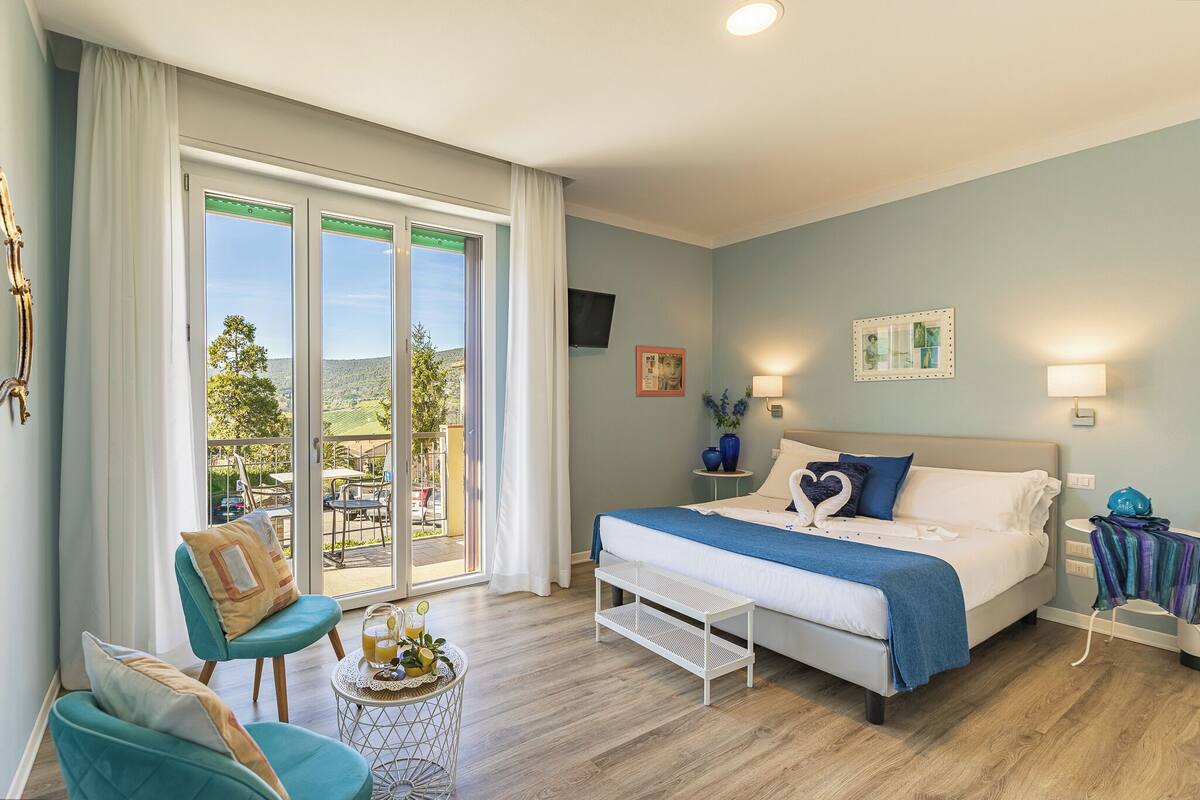 A bedroom with a large bed, a chair, a small table with a drink, and a view of the outdoors through a sliding door.