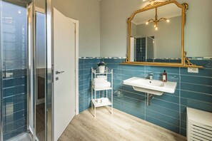 Un baño con un lavamanos blanco, un espejo con marco dorado y un estante blanco con artículos de aseo.