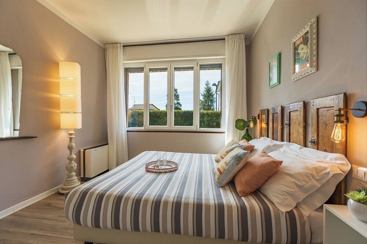 A bedroom with a large window, a bedside table with a lamp, and a bed with striped bedding.