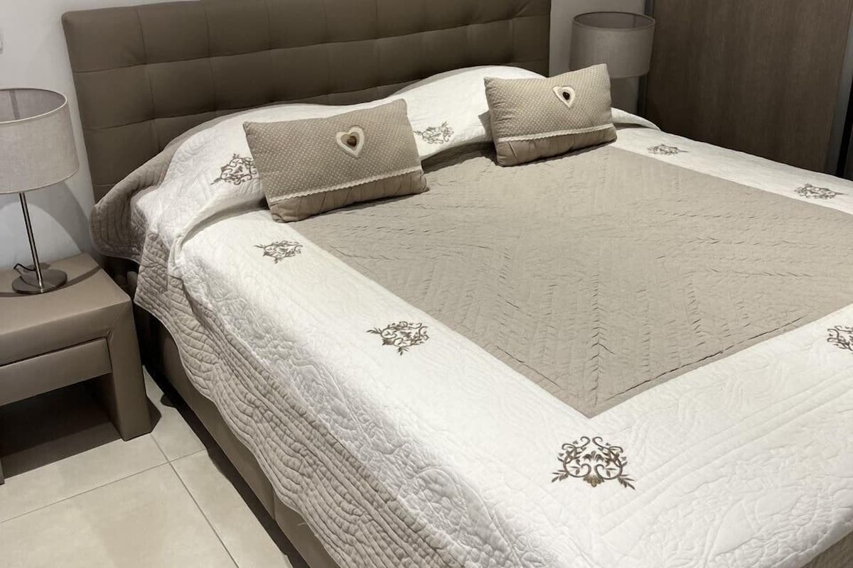 Un lit bien fait, recouvert d’une courtepointe blanche matelassée et agrémenté de coussins décoratifs, une lampe de chevet et un tableau de fleurs accroché au mur.