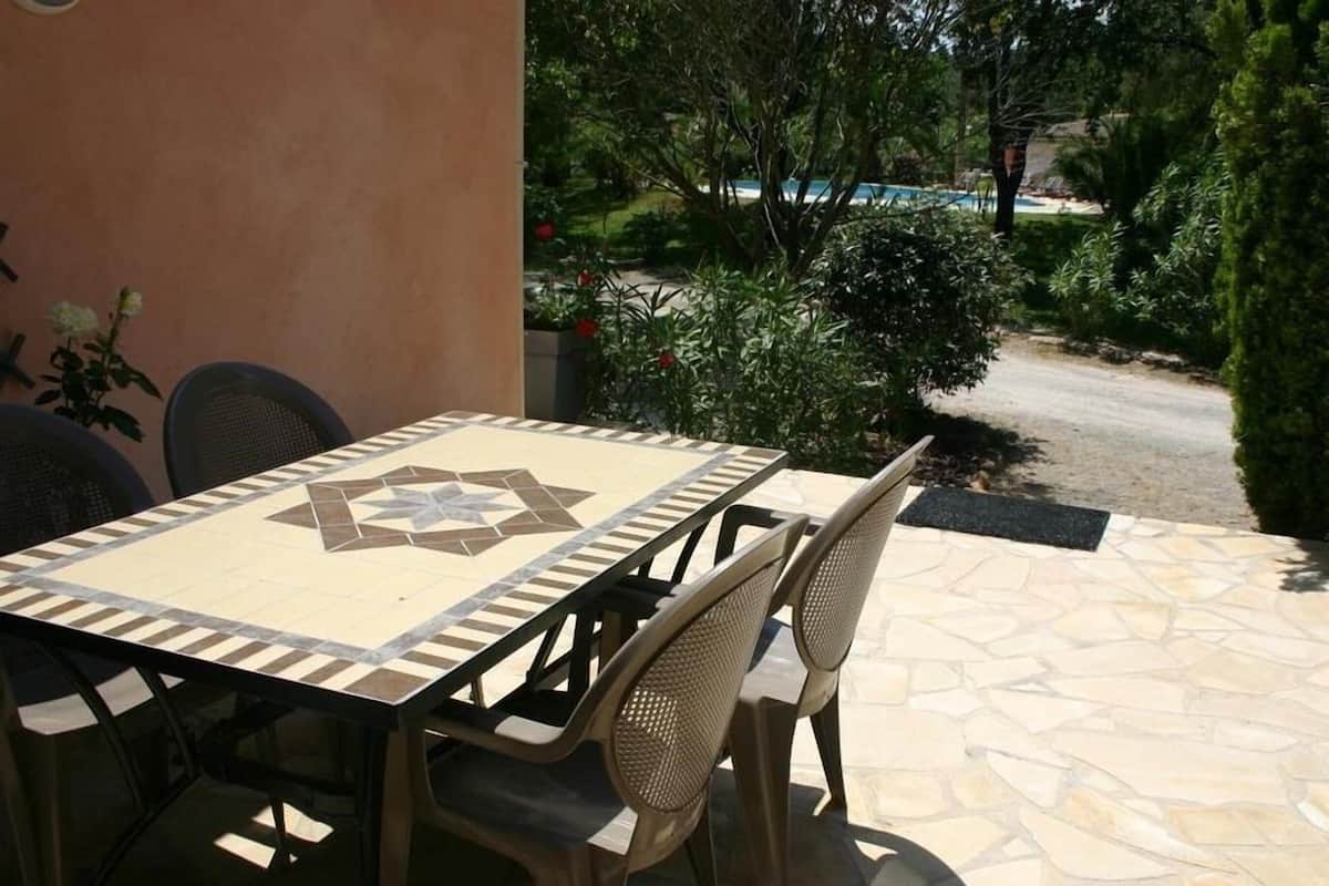 Une terrasse extérieure avec une table et des chaises carrelées, entourée de verdure et d’une piscine.