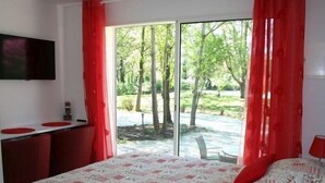 Quarto com uma cama grande, TV fixada na parede e porta de vidro deslizante para área externa com árvores.