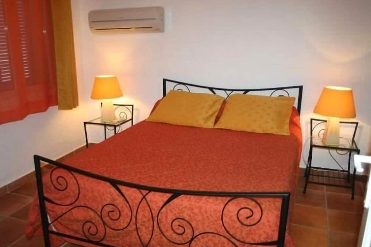 Un lit simple avec une couvre-lit rouge et deux oreillers, flanqué de deux tables de chevet avec des lampes, dans une pièce dont le sol est carrelé et où se trouve un climatiseur mural.