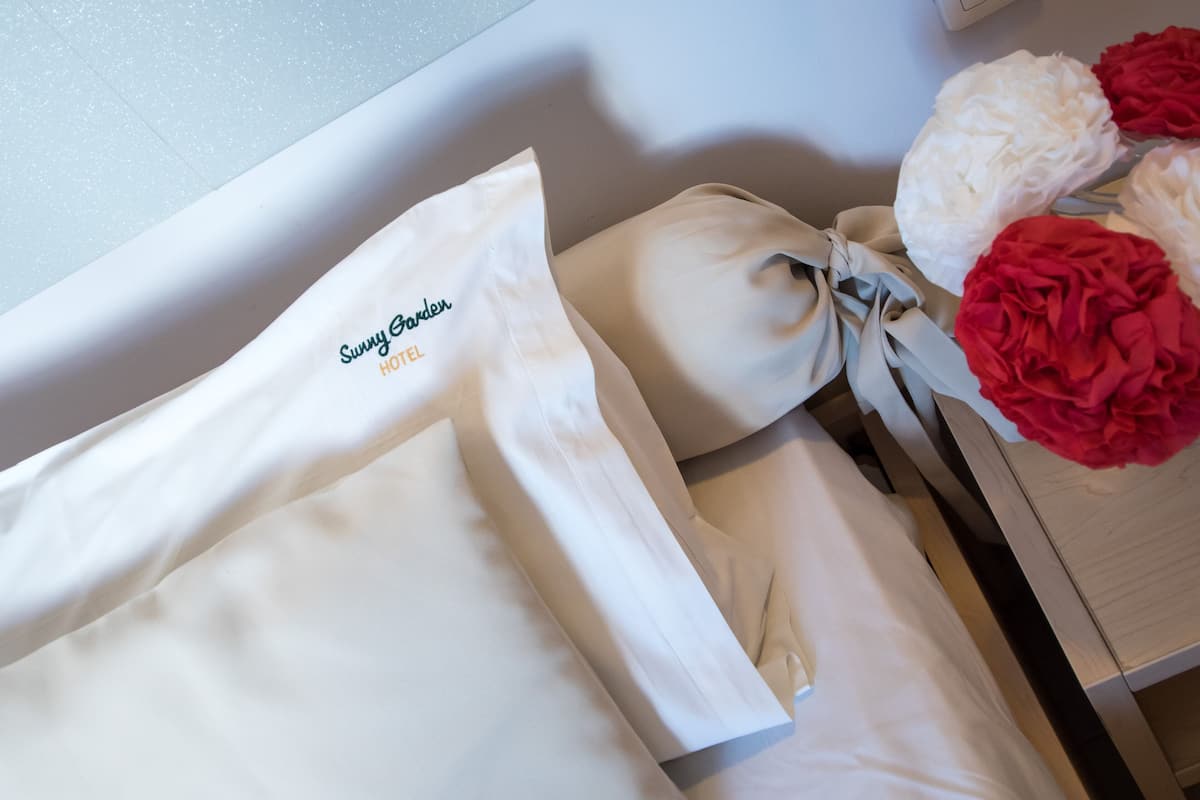 En prydligt vikt, vit hotellhandduk med broderad text som lyder ”Sunny Garden Hotel”, placerad på en träyta bredvid en säng dekorerad med vita och röda blommor.