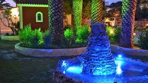 A stone fountain with water features, surrounded by palm trees and a building with a red facade. - Sunny Garden (Zakynthos)