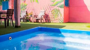 A swimming pool with a pink wall featuring a tropical leaf mural, a covered seating area, and a building in the background.