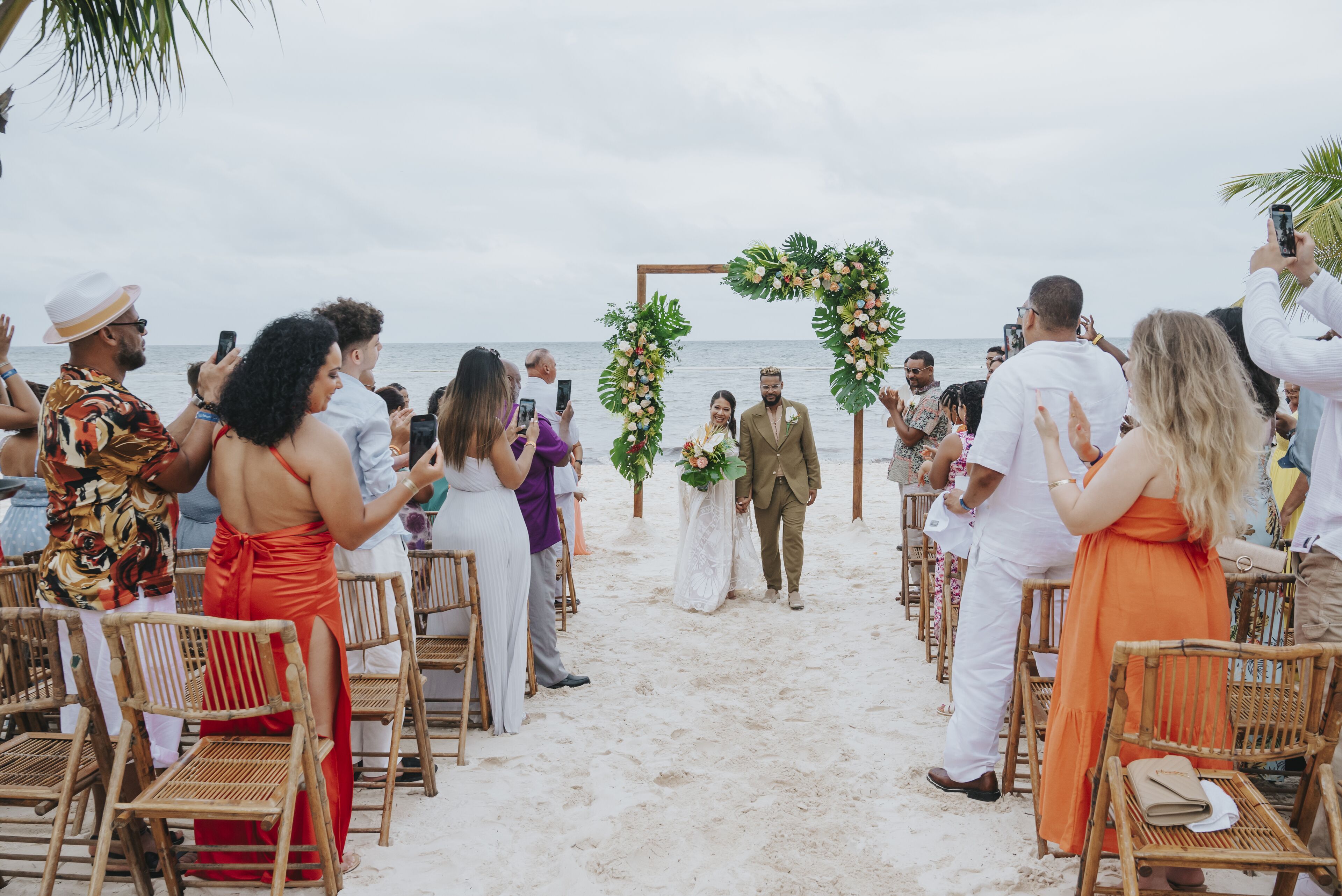 Una cerimonia nuziale sulla spiaggia con invitati seduti su sedie in vimini, un arco floreale e una spiaggia sabbiosa sullo sfondo.
