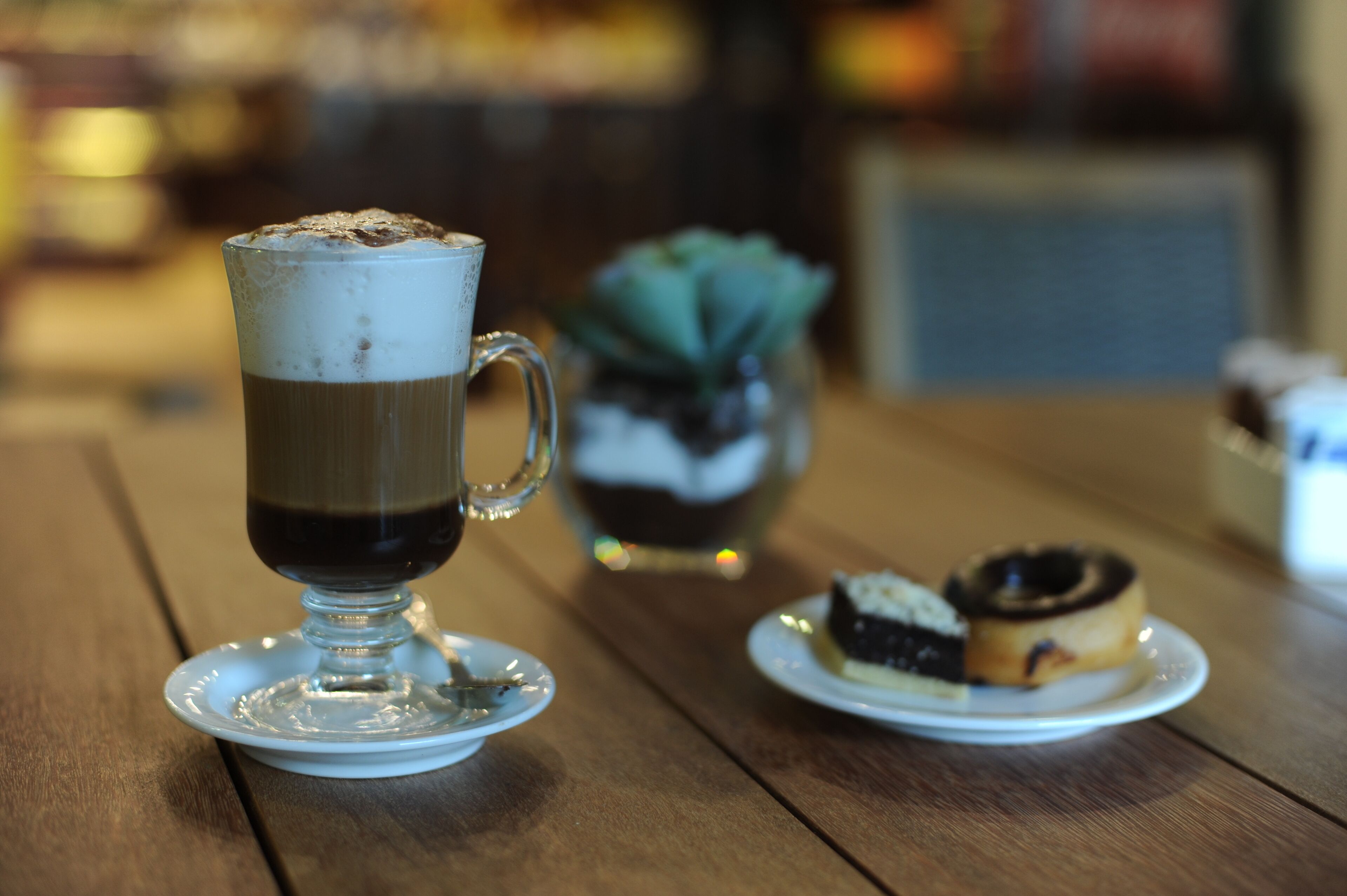 A glass of coffee on a saucer, a small plate with a pastry, and a potted plant on a wooden table.