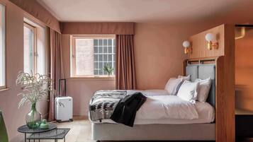 A bedroom with a bed, a suitcase, a green chair, a small table with a vase, and a window with curtains.