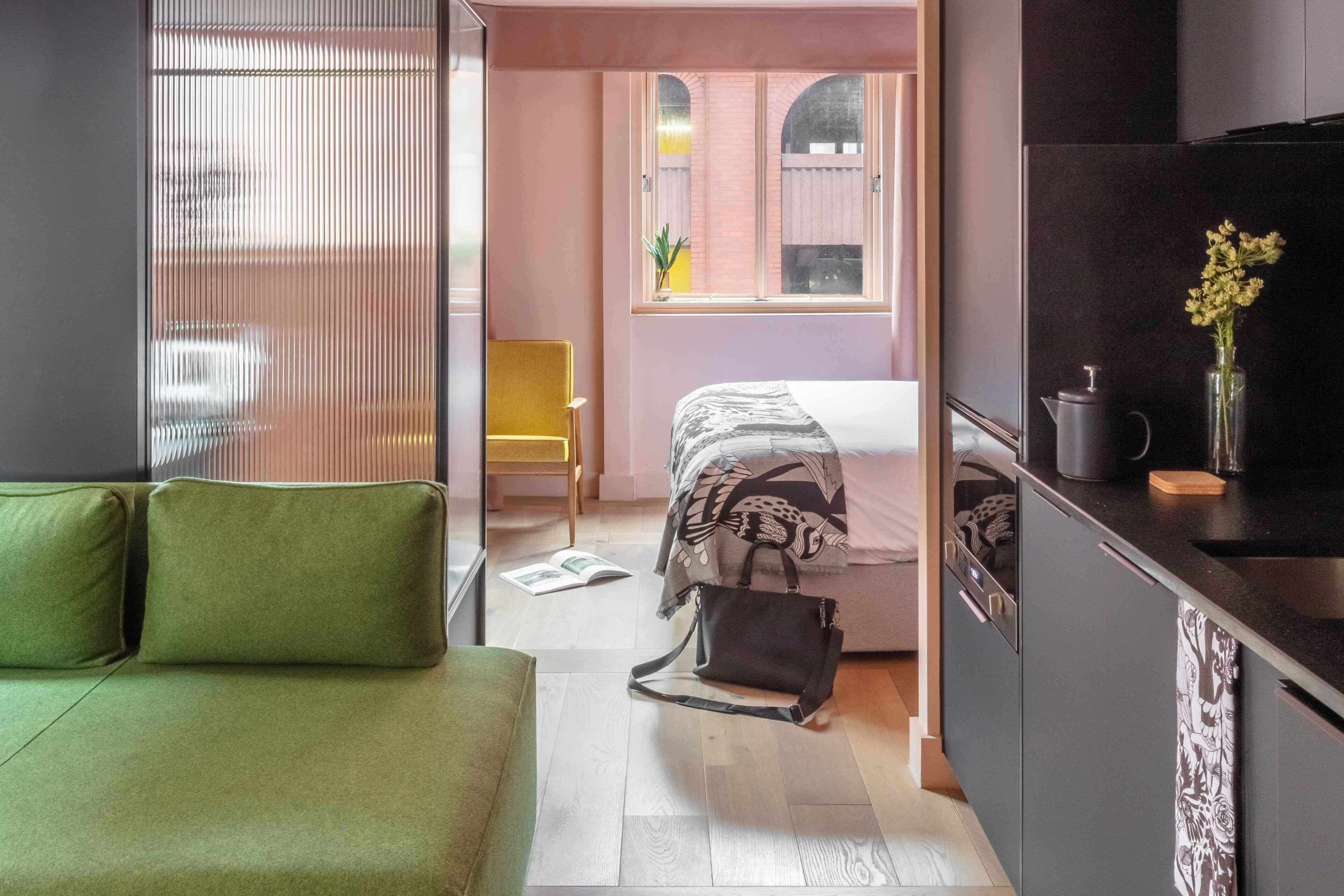 A compact hotel room with a green sofa, a bed with patterned bedding, a small kitchen area, and a view through a window.