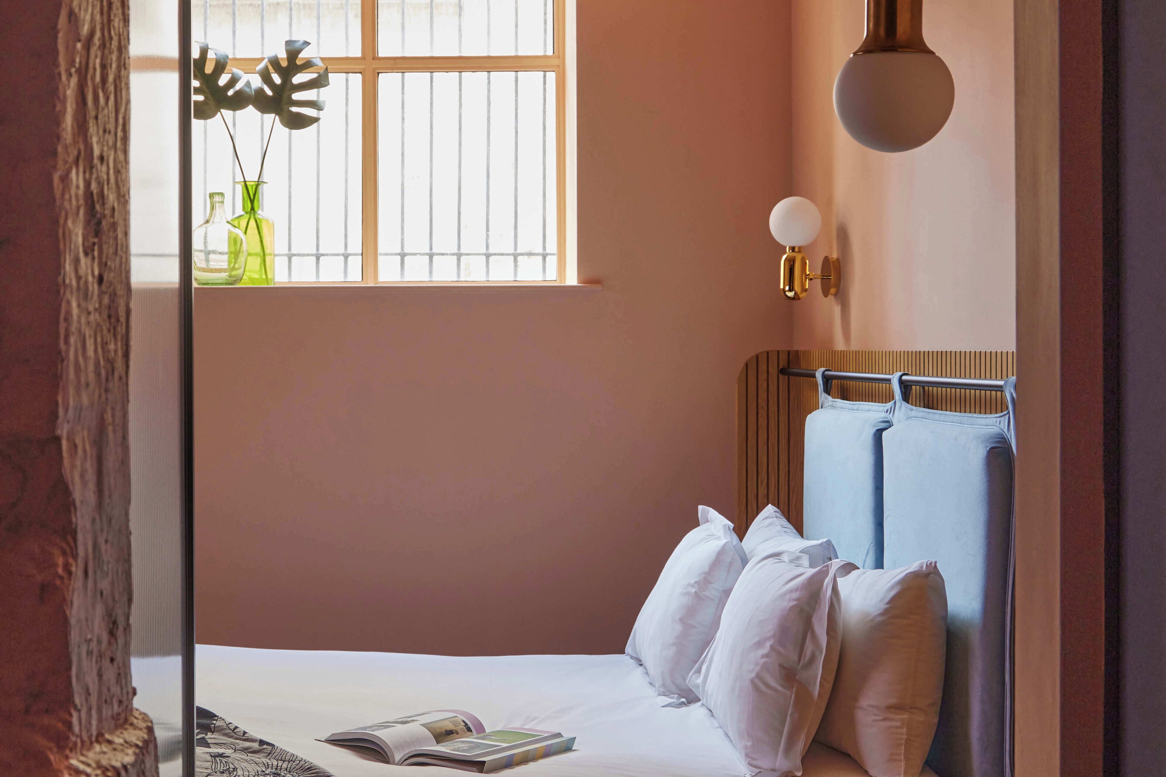 Un dormitorio con cama, almohadas, una ventana con rejas, una lámpara de pared y un cabecero de madera.