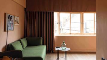 A room with a green sofa, a small round table with a bottle, a chair, and a window with curtains.