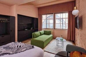 Une chambre d’hôtel moderne avec un canapé vert, un lit recouvert d’une couverture à motifs, une petite table sur laquelle est posé un vase, et une grande fenêtre donnant sur un immeuble.